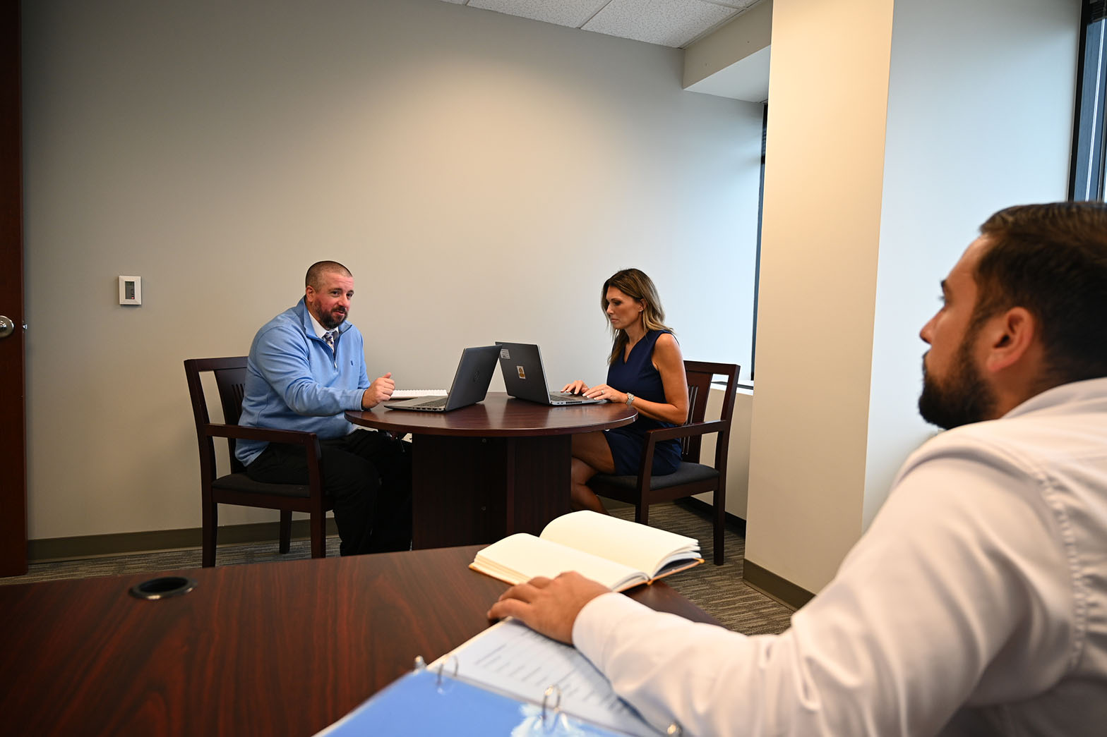 Three colleagues in an office meeting: two on laptops at a round table, one reviewing an open binder.